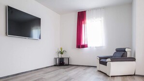 A modern living room with a flat-screen TV mounted on the wall, a comfortable sofa, a small table with a potted plant, and a window with red curtains. - Holiday Home J&D No.3-Six Bedroom House  with Pool (Podstrana)