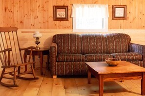 Una habitación acogedora con piso de madera, un sofá estampado, un sillón mecedor, una mesita con lámpara y una ventana con cortinas.