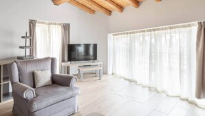 Un salon avec un canapé gris, un téléviseur, une table basse en bois et une fenêtre ornée de rideaux légers.