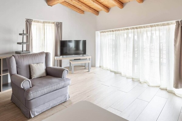 A living room with a grey sofa, a TV, a wooden coffee table, and a window with sheer curtains.