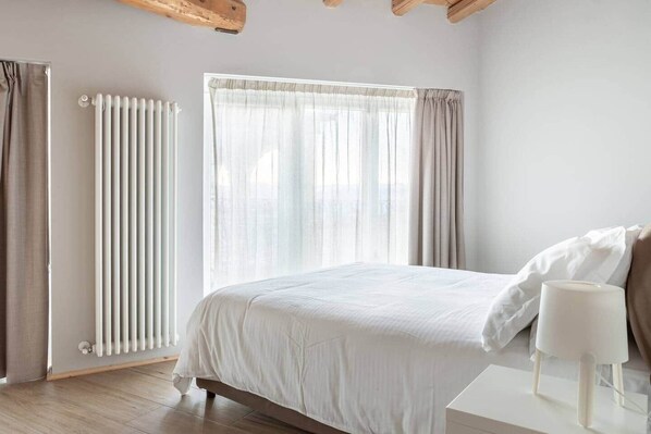Un dormitorio con cama, mesita de noche, radiador y ventana con cortinas.