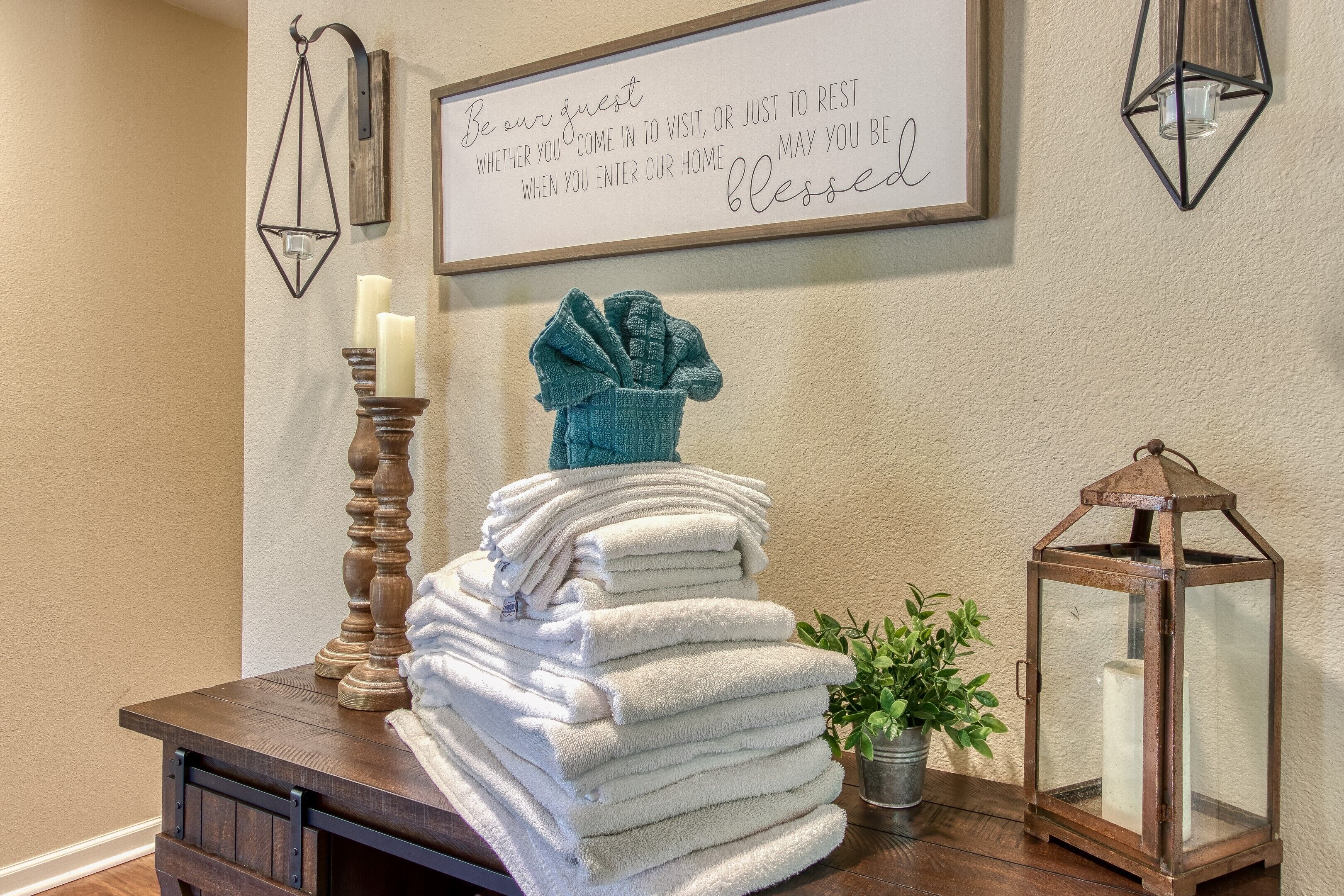 A stack of white towels on a wooden table, a teal towel on top, a candle holder with candles, a lantern, and a framed sign on the wall.