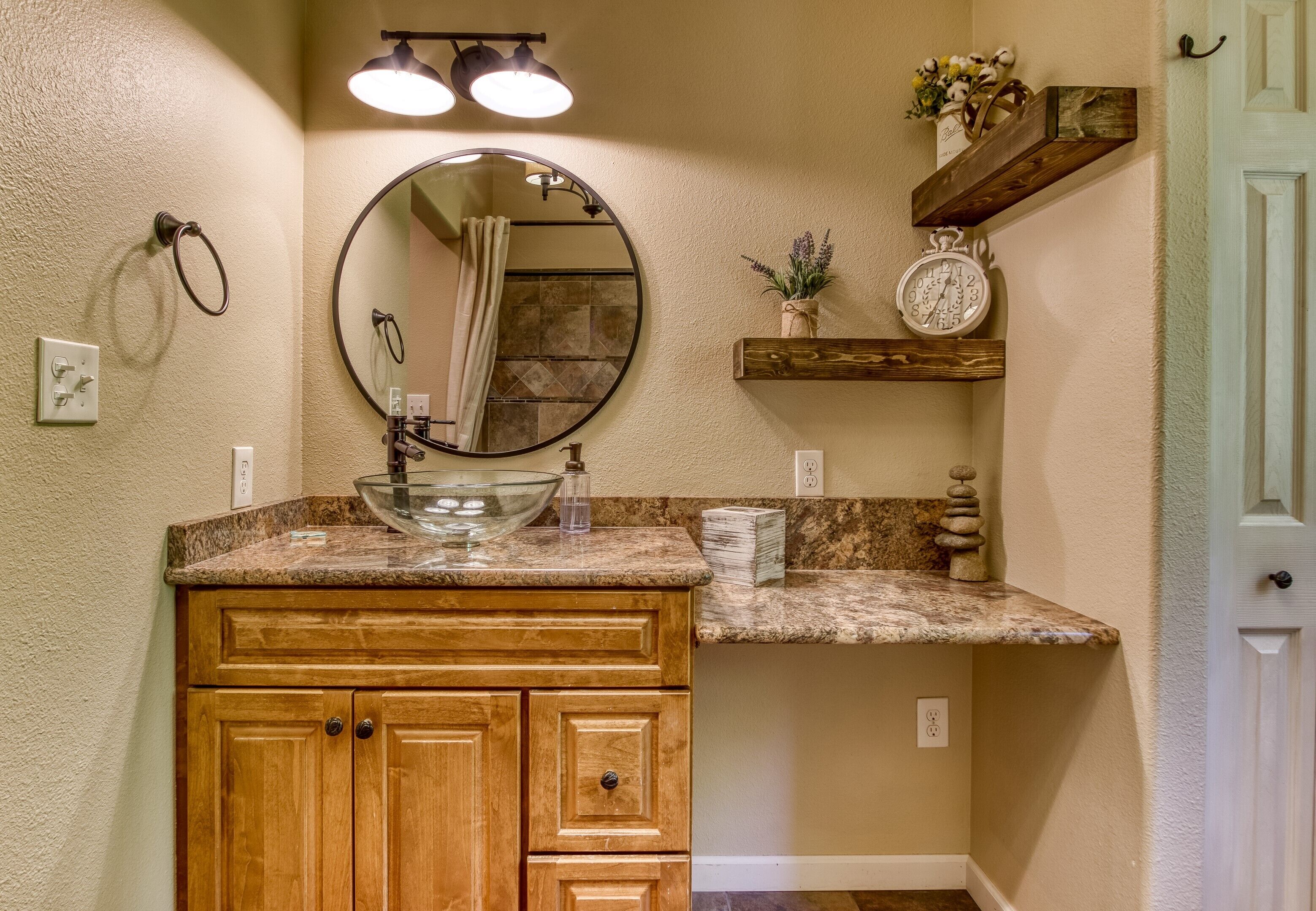 Une salle de bain avec un meuble-lavabo en bois, un miroir rond, un lavabo en verre en forme de bol, une horloge et une étagère ornée d’objets décoratifs.