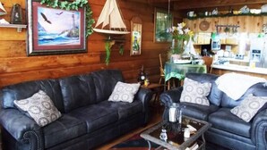 Ein gemütliches Blockhauss-Interieur mit einem dunkelbraunen Ledersofa, einem kleinen Couchtisch und einem durchgehenden Durchgang zur Küche.