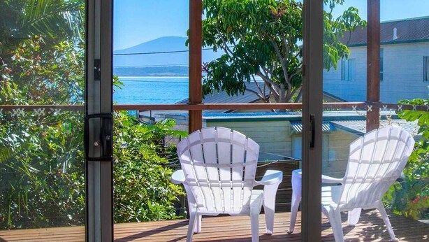 A covered outdoor area with two white chairs, a wooden deck, and a view of the ocean and a mountain.