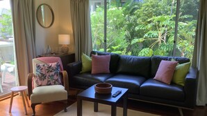 A living room with a dark leather sofa, a small table, a chair, and a view of lush greenery outside.