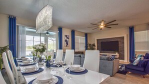 Uma sala de jantar com uma mesa branca posta para seis pessoas, jogos americanos azuis e cadeiras brancas. Há um lustre sobre a mesa e um ventilador de teto na sala de estar.