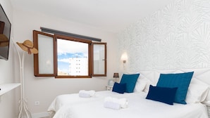 Ein Schlafzimmer mit zwei Betten, weißer Bettwäsche, blauen Kissen, einem weißen Kopfteil, einem Nachttisch mit Lampe, einem Spiegel und Blick auf Gebäude durch ein Fenster.