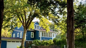 A two-story house with a garage, surrounded by trees and a stone wall. - Woodstock Farmhouse /Hot tub, Gardens,EV charger, Firepit, Pet/Family Friendly (Saugerties)