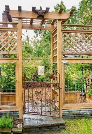 Una estructura de pérgola de madera con un adorno en forma de estrella, un portón metálico con detalles decorativos, y un jardín con diversas plantas.