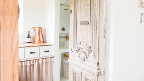 Een houten deur in landelijke stijl met een schuifmechanisme, een witte muur, een klein houten bankje, een koperen waterkoker op een aanrecht en een vloerkleed met dessin.