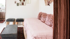 Een slaapkamer met een bed, twee krukken, een vloerkleed en diverse wanddecoraties, waaronder spiegels en een veer.