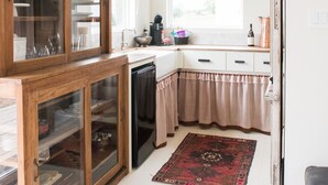 Een keuken met een houten kast, een vaatwasser, een raam met zonwering, een hanglamp en een rood vloerkleed.