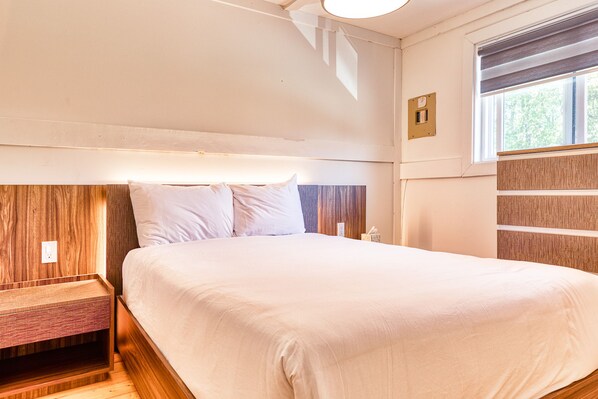 A neatly made bed with a wooden headboard, a bedside table, and a window with blinds.