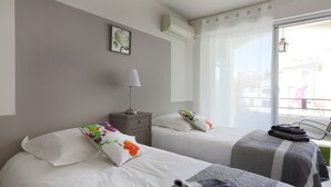 A hotel room with two beds, a bedside table with a lamp, a window with curtains, and a view of the outside.