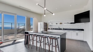 Une cuisine moderne dotée d’un grand îlot central, de tabourets de bar et offrant une vue sur l’océan grâce à de grandes portes-fenêtres coulissantes.