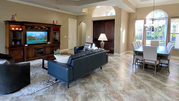 Un salón espacioso con un televisor de pantalla plana grande, un sofá azul oscuro, una zona de comedor con mesa y sillas, y vistas al exterior a través de amplios ventanales.
