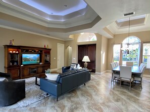 Un salón espacioso con un televisor de pantalla plana grande, un sofá azul oscuro, una zona de comedor con mesa y sillas, y vistas al exterior a través de amplios ventanales.