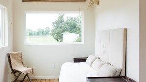 Un dormitorio con suelo de madera, una cama con ropa de cama blanca, una silla, una ventana con vistas a árboles y una lámpara fijada a la pared.