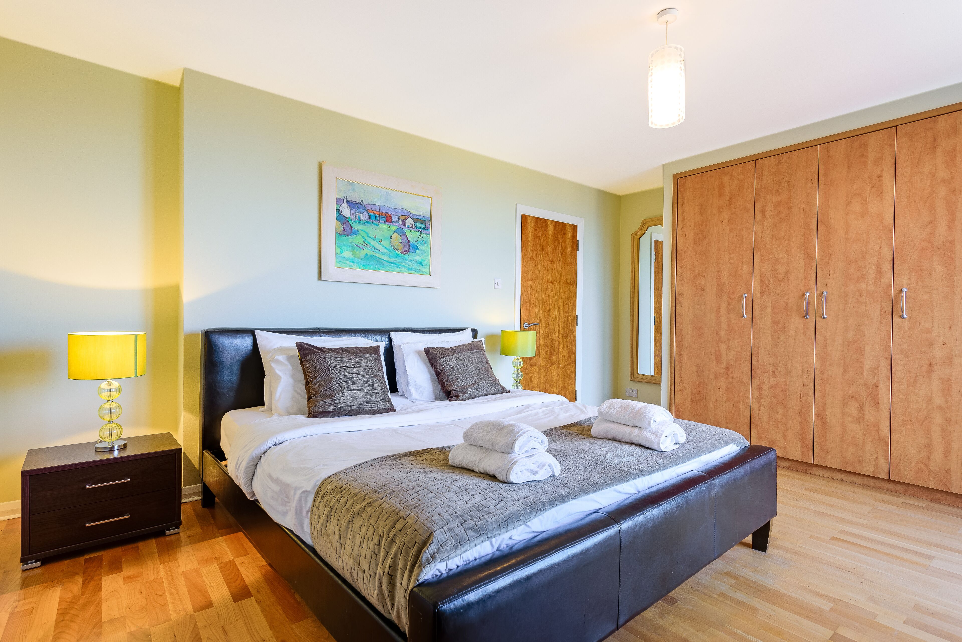 A well-lit bedroom with a large bed, bedside lamps, a wooden wardrobe, and a painting on the wall.