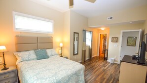A bedroom with a bed, nightstands, a ceiling fan, a window with blinds, a door, and a small desk with a television.