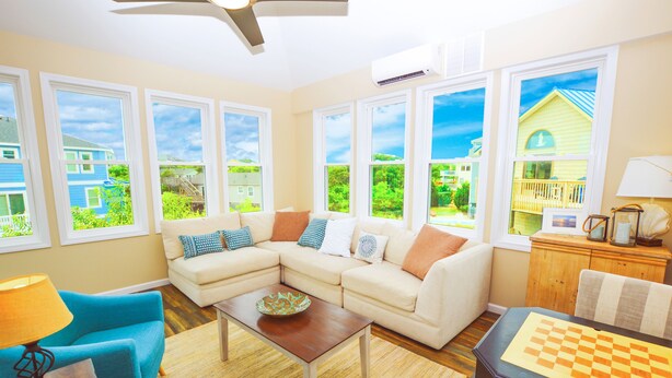 A living room with a sectional sofa, a coffee table, a chessboard, and large windows offering a view of houses and greenery.