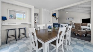 A dining area with a wooden table and white chairs, a kitchen in the background, and a ceiling fan.