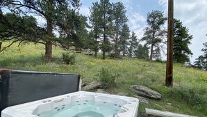 A hot tub in an outdoor setting with a wooden bench nearby, surrounded by trees and a grassy area with yellow flowers. - The Rose Den - Romantic Mountain Cabin with Views! (Estes Park)