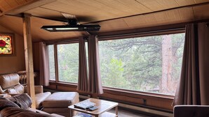 A cozy room with a large window, a ceiling fan, a leather sofa, and a small table with a book. - The Rose Den - Romantic Mountain Cabin with Views! (Estes Park)