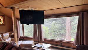 A cozy living room with a leather couch, a coffee table, and a flat-screen TV. Large windows offer a view of trees and a forest. - The Rose Den - Romantic Mountain Cabin with Views! (Estes Park)