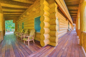 Una cabaña de troncos con un porche de madera, varias sillas Adirondack y un ventanal con vistas a la vegetación.