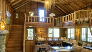 A rustic cabin interior with a wooden staircase, a dining area, and a living room with leather sofas and a round coffee table.