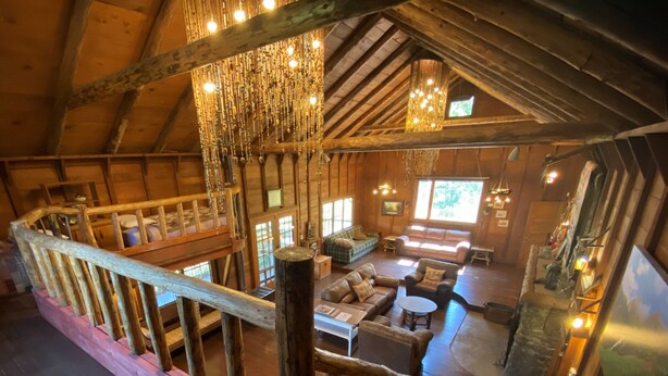 A rustic cabin interior with a loft bedroom, wooden beams, a wooden staircase, and a cozy living area with sofas and a fireplace.