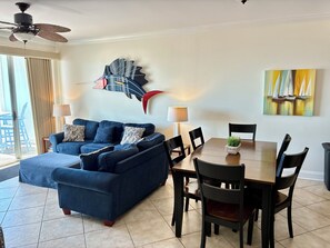 A living room with a sofa, dining table, chairs, a ceiling fan, and a wall art piece.
