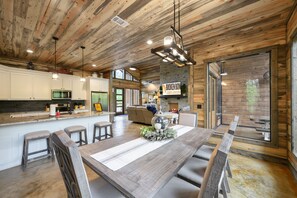 A rustic kitchen with wooden beams, a large island with stools, and a stone accent wall. - Shady Creek Lodge- 5 Bedroom, 4 kings plus bunk room! (Broken Bow)