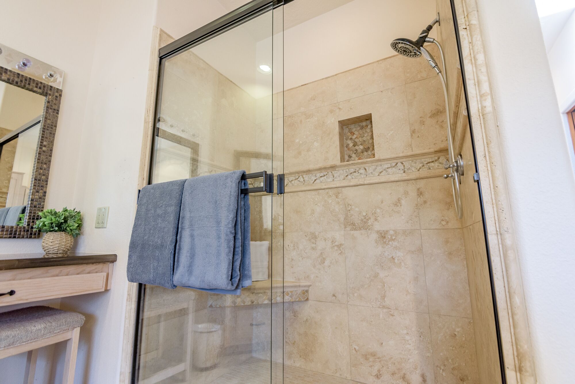 A glass-enclosed shower with a handheld showerhead, a towel rack with two towels, a wooden vanity with a plant, and a mirror with a decorative frame.