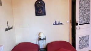 A room with two single beds, a small table with a lamp, a door with a decorative panel, and wall decorations including a clock and a framed certificate. - Large studio located opposite the spa park. (Vittel)
