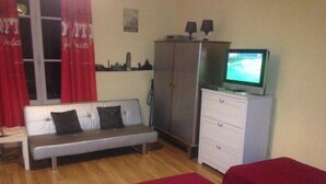 A room with a sofa, a television on a cabinet, a wardrobe, and a bed with red bedding. - Large studio located opposite the spa park. (Vittel)