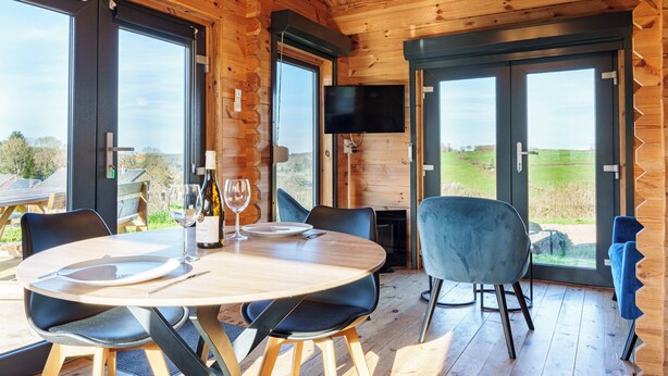 L’intérieur d’un chalet en bois présente une table à manger dressée pour deux personnes, un téléviseur fixé au mur et de grandes fenêtres offrant une vue sur l’extérieur.
