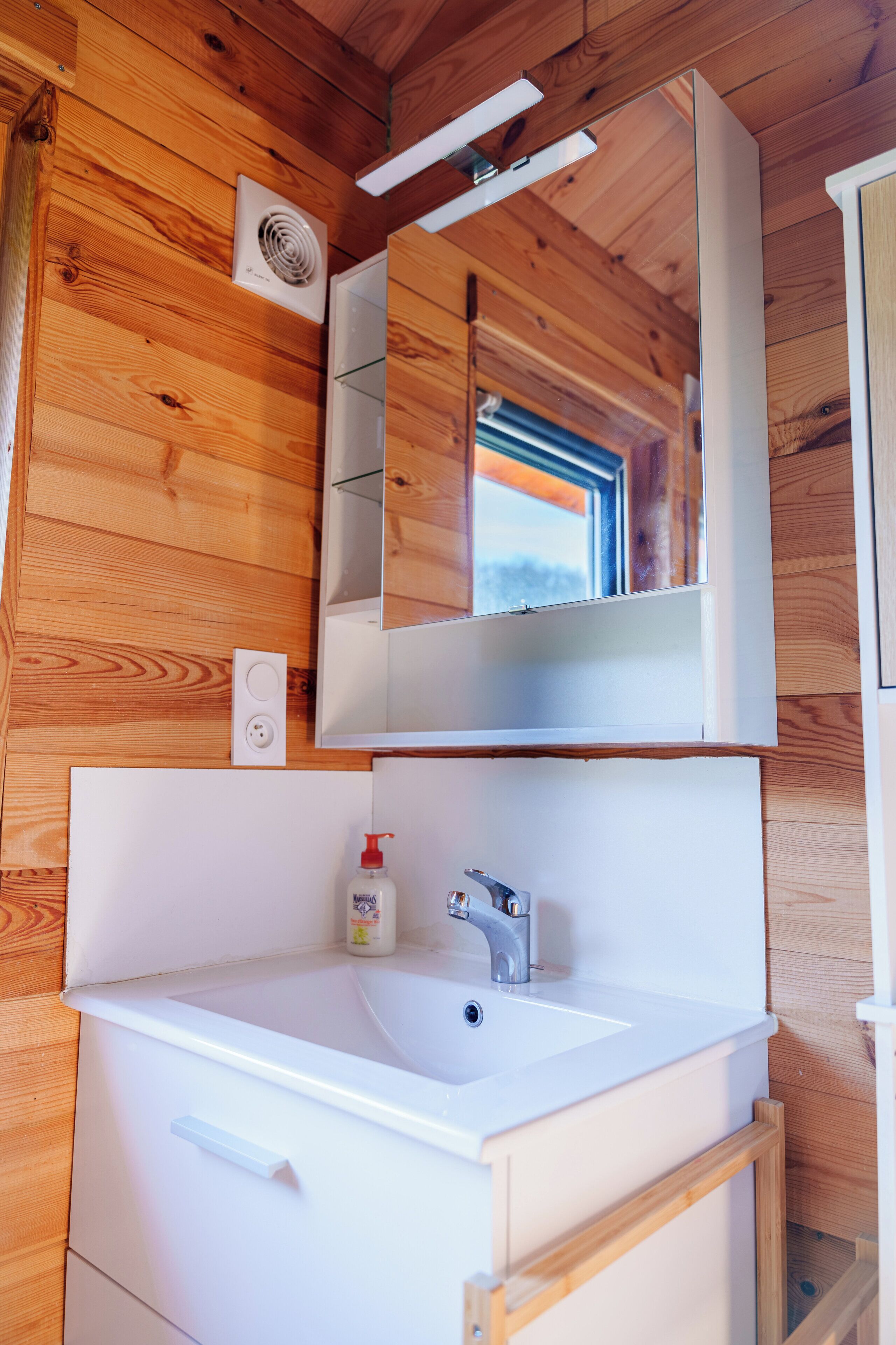 A wooden cabin interior with a white sink, a mirror above it, and a window with a view of the outdoors.