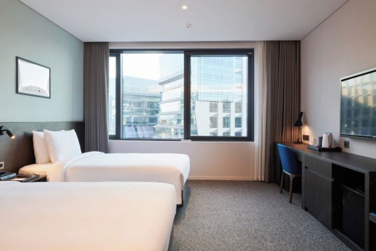 A hotel room with two beds, a desk, a chair, a TV, and a large window with a city view.