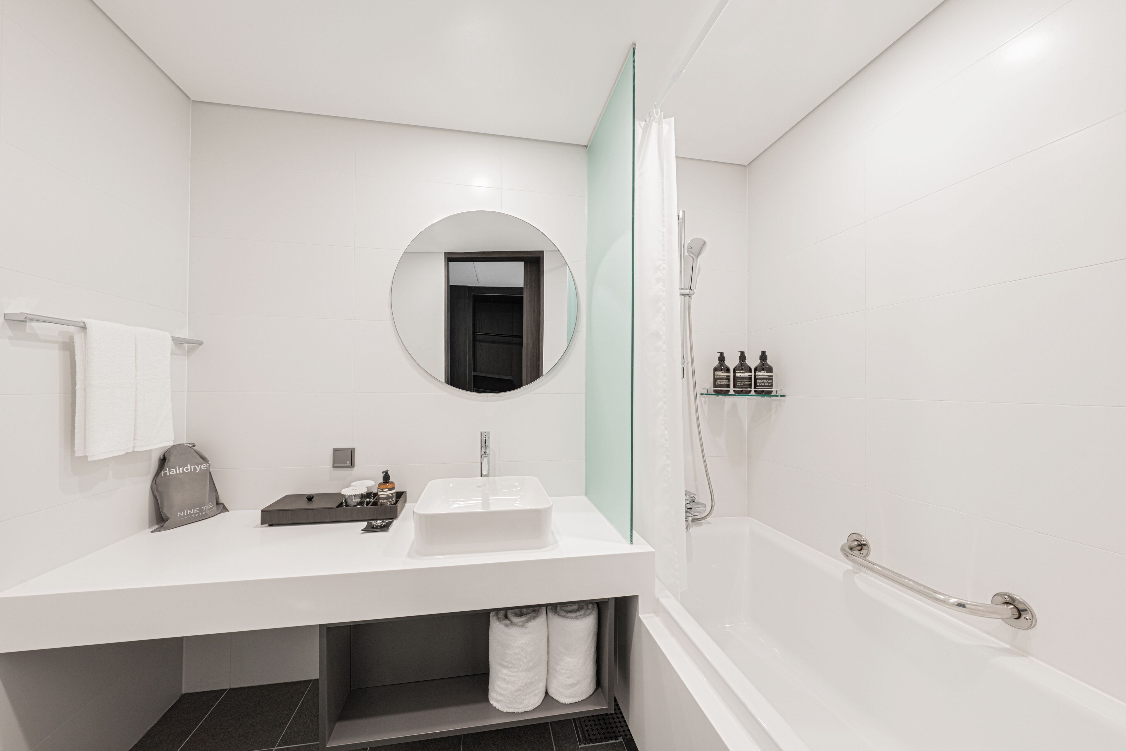 A modern bathroom with a white vanity, a round mirror, a glass shower enclosure, and a bathtub with a grab bar.