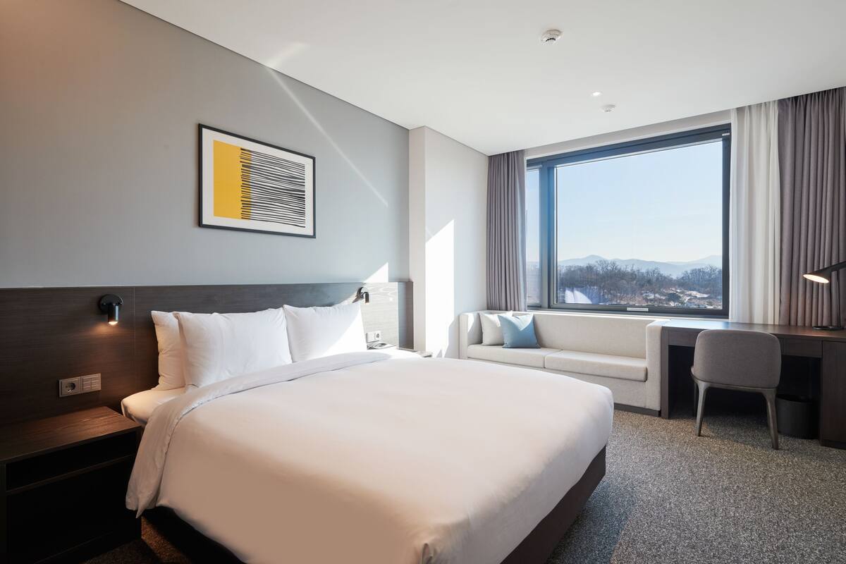 Ein Hotelzimmer mit einem großen Bett, einem Schreibtisch und Blick auf die Berge durch ein großes Fenster.