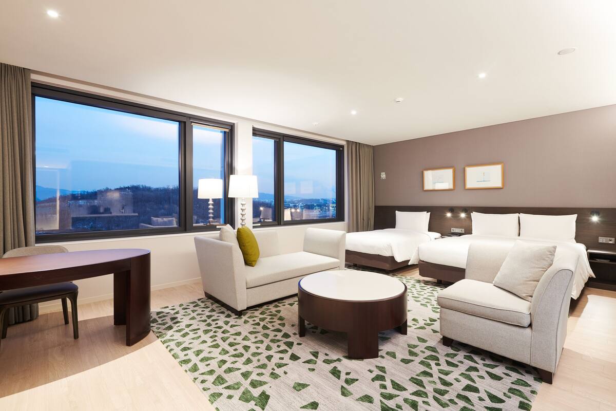 Ein modernes Hotelzimmer mit großem Fenster, einer Couch, einem Bett, einem runden Couchtisch und einem kleinen Schreibtisch.