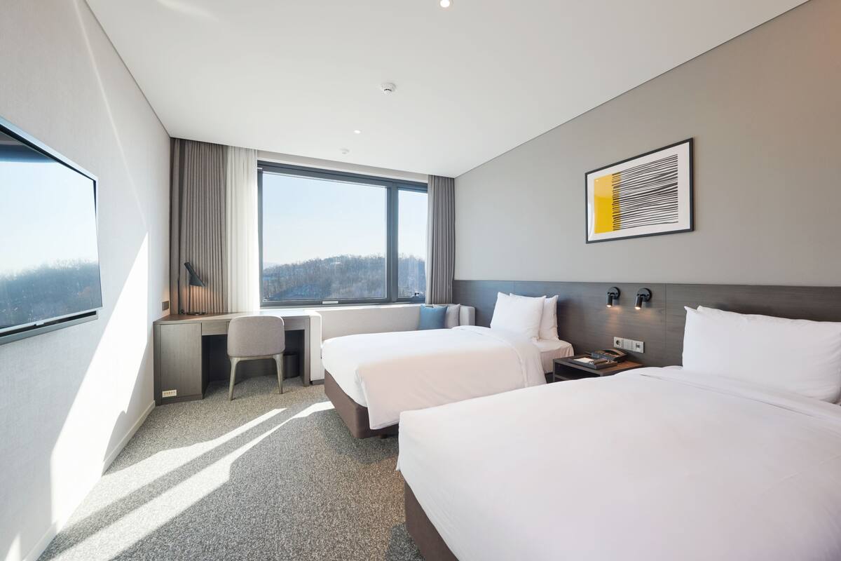 Ein modernes Hotelzimmer mit zwei Betten, einem Schreibtisch und einem großen Fenster mit Blick auf die Stadt.