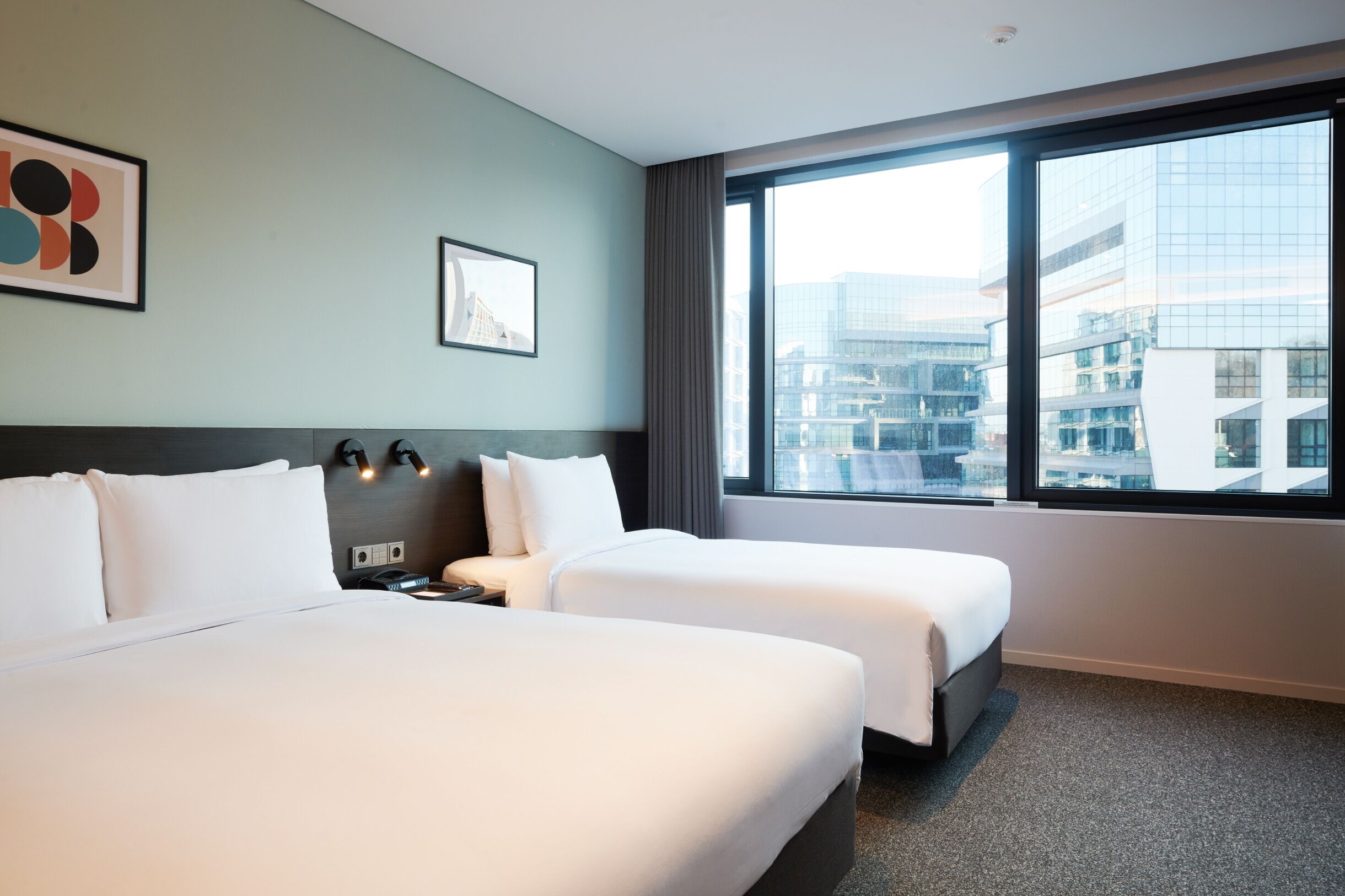 A hotel room with two beds, a large window with a city view, and artwork on the walls.
