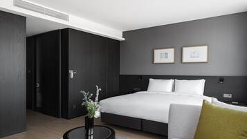 A modern hotel room with a large bed, a bedside table with a vase, a grey sofa, and two framed pictures on the wall.