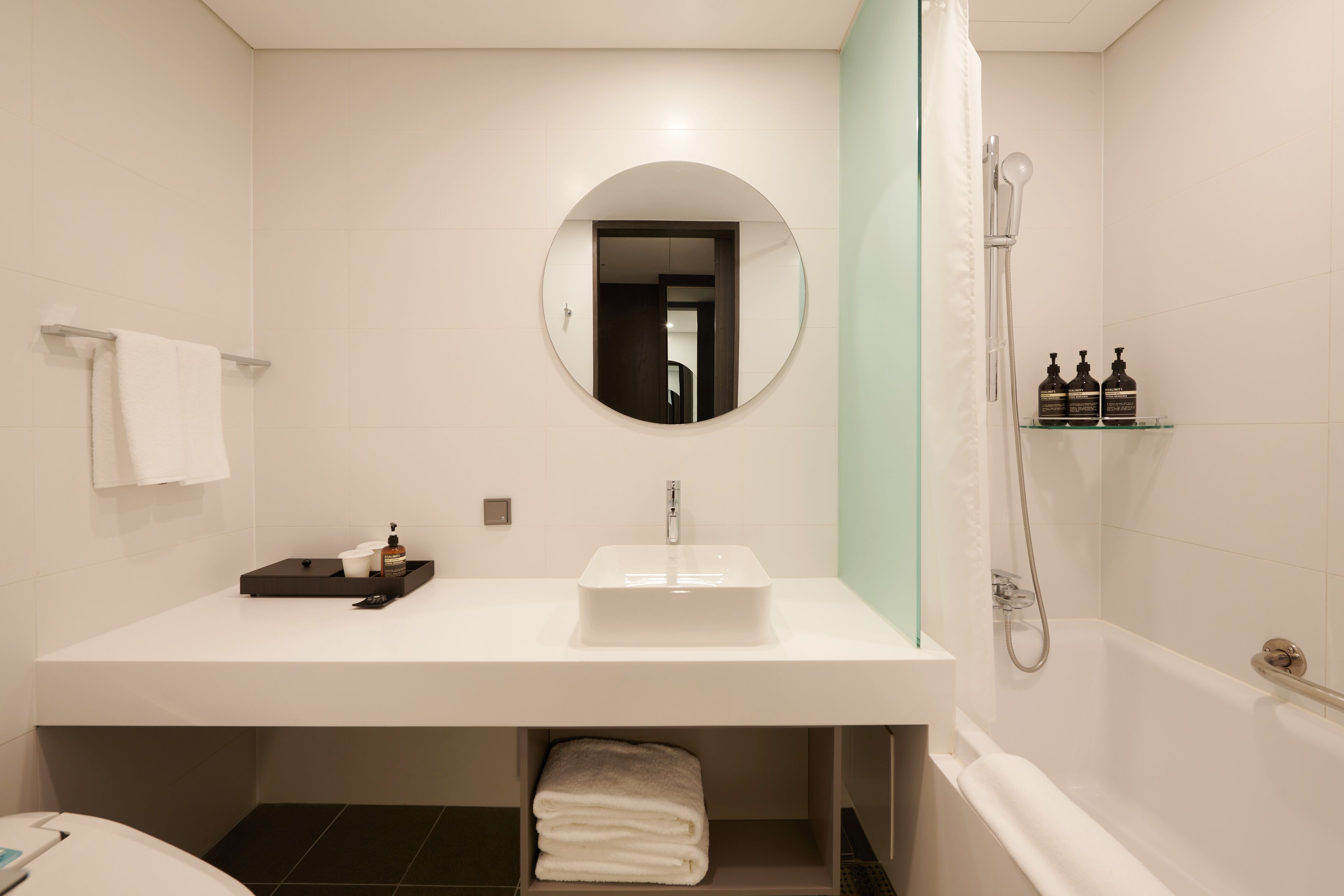 A modern bathroom with a white sink, a round mirror, a shower, and a glass shelf with toiletries.
