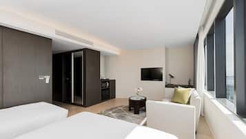 Una habitación de hotel moderna con dos camas, un sofá, una mesita con un florero, un televisor y un ventanal grande.
