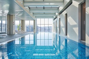 Una piscina interior con paredes de vidrio transparente, sillas de descanso y vistas panorámicas de la ciudad.